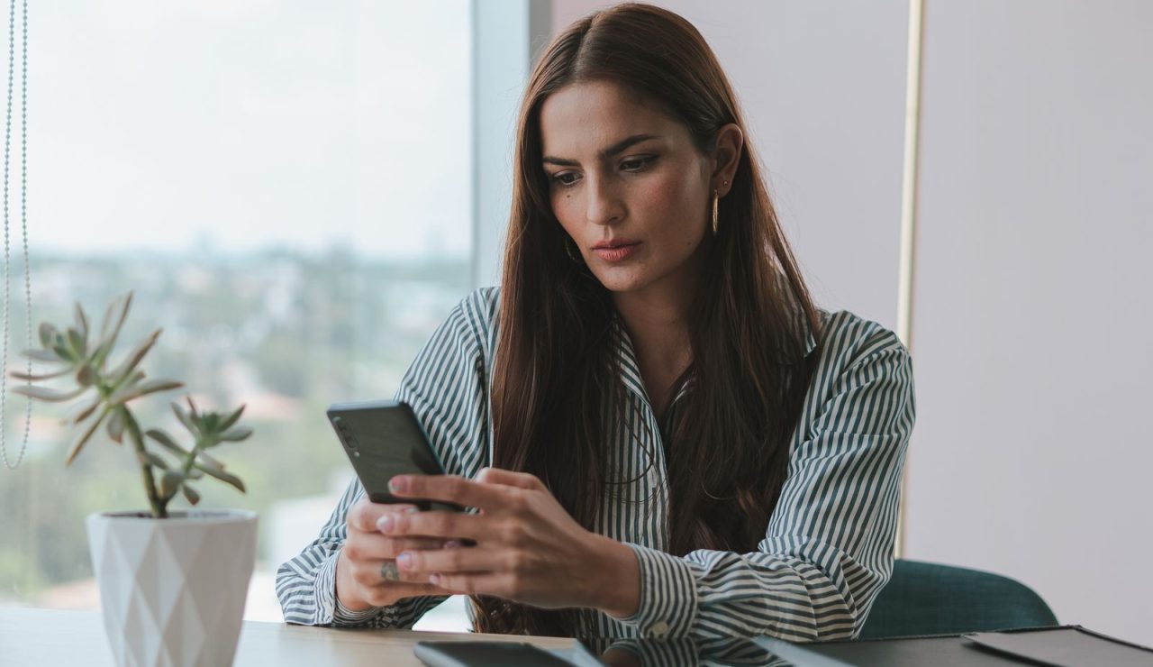 Jeune candidate assise à un bureau devant une baie vitrée, consultant son téléphone d'un air concentré