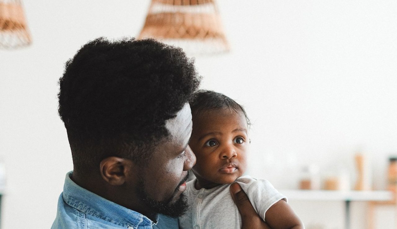 Un pere porte son bebe dans une cuisine lumineuse, illustration du conge supplementaire de naissance ouvert aux parents salaries
