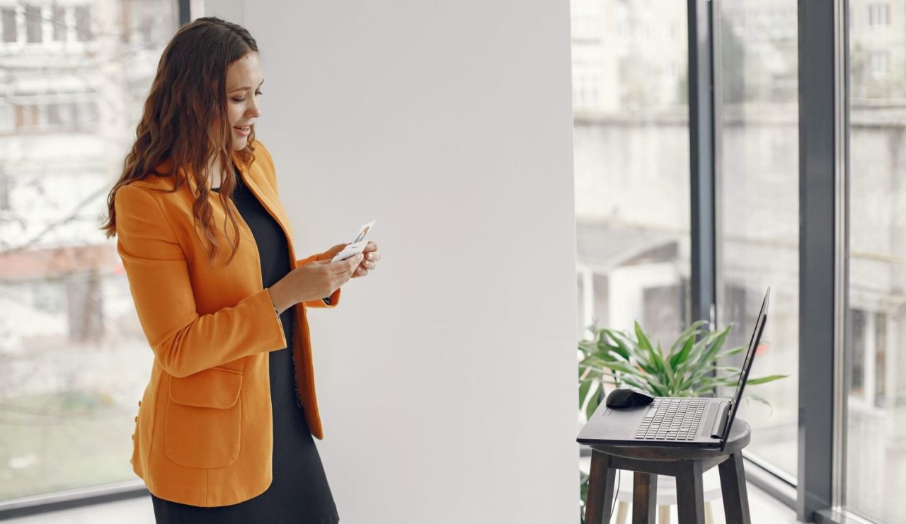 Cadre en veste orange consultant son téléphone dans un bureau vitré lumineux avant un entretien de présélection