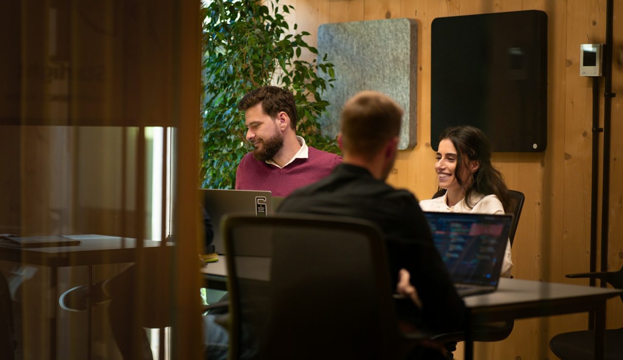 Deux collègues discutent avec le sourire autour d'un ordinateur portable dans un bureau lumineux en bois clair