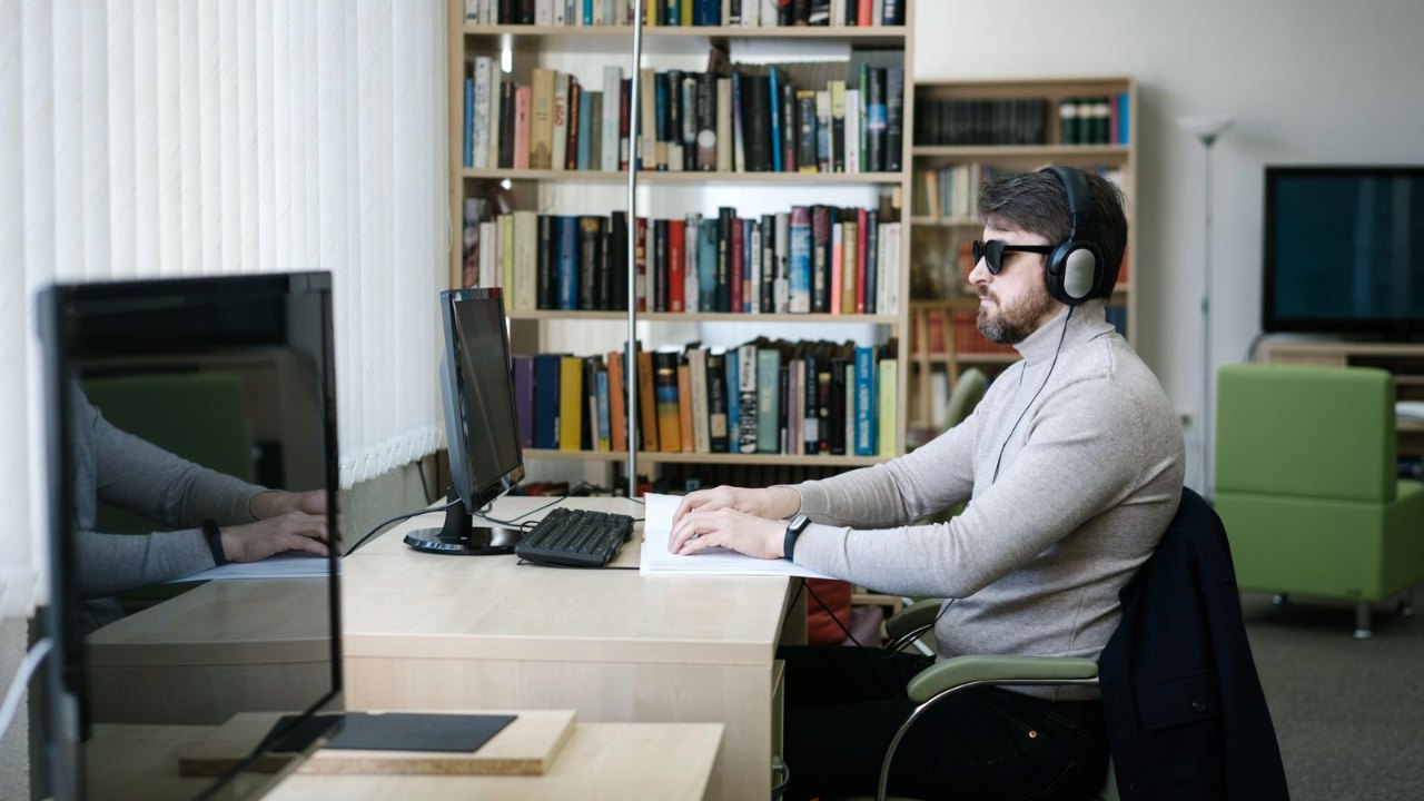 Un homme malvoyant équipé d'un casque audio travaille à un poste informatique doté d'une plage braille dans un bureau lumineux