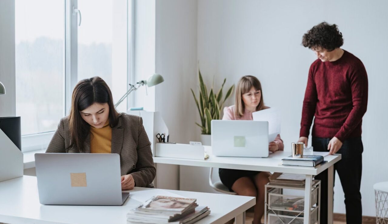 Une salariee travaille sur son ordinateur portable dans un bureau lumineux, deux collegues echangent en arriere plan