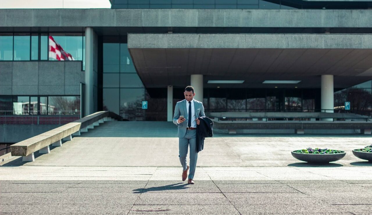 Un salarié en costume gris quitte un immeuble de bureaux en béton, veste sur le bras, en fin de semaine